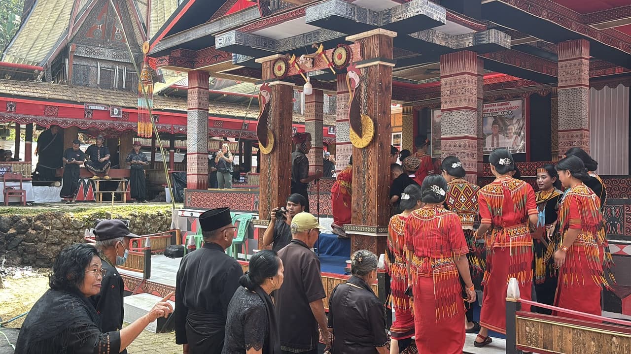 A group of guests in red traditional dress with fringed sashes arriving at a decorated pavilion, older men in black nearby.