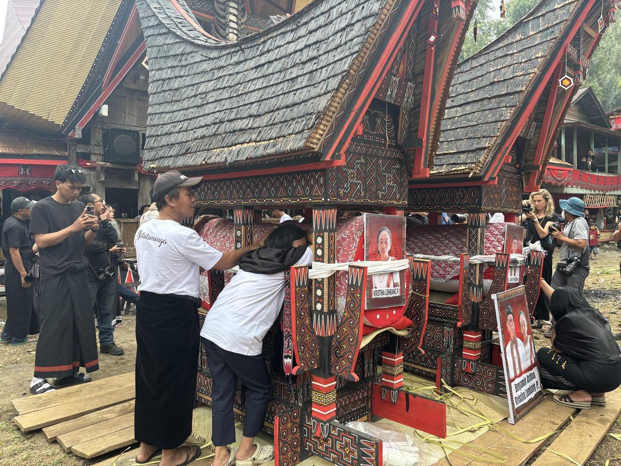Family members leaning over the coffins on the bier, crying, photo cards reading "Kristina Lembang P." and "Benyamin Anthon" visible.