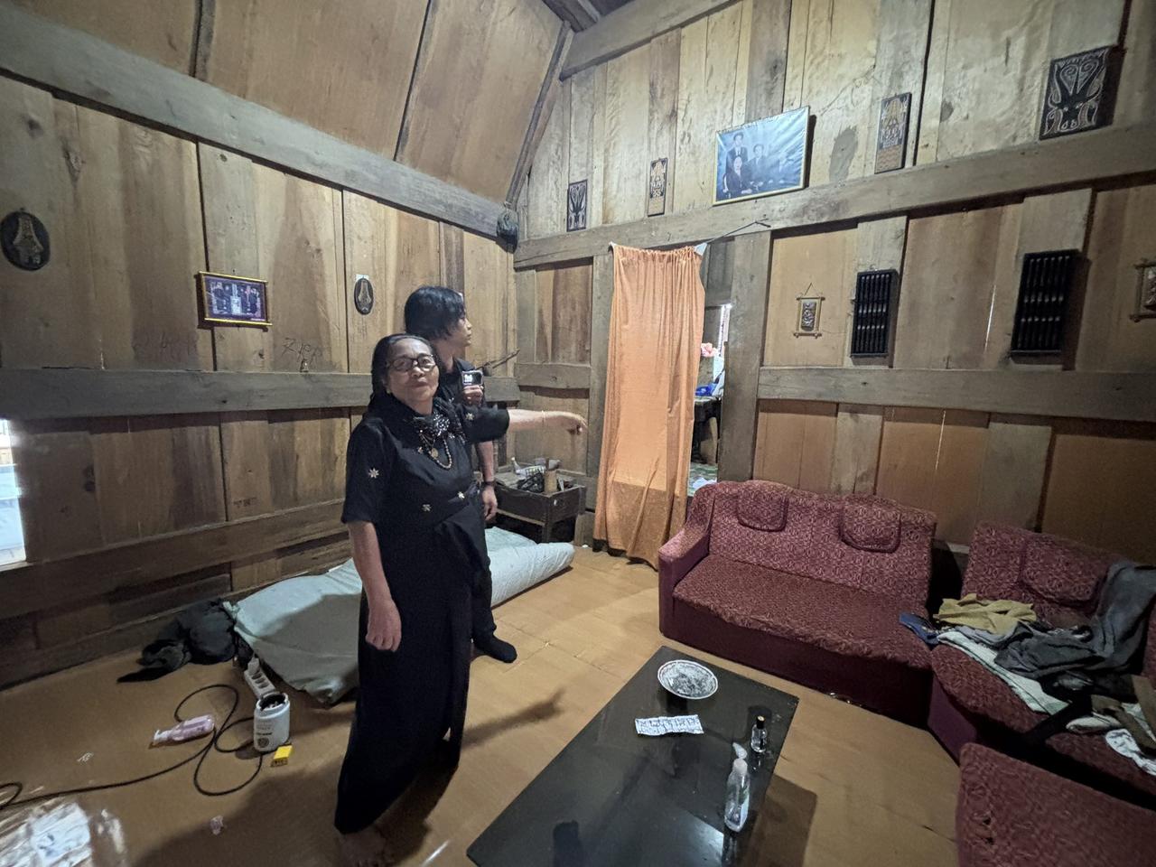 Family members gathered inside a dim wood-paneled room of the tongkonan, photographs on the walls above them.