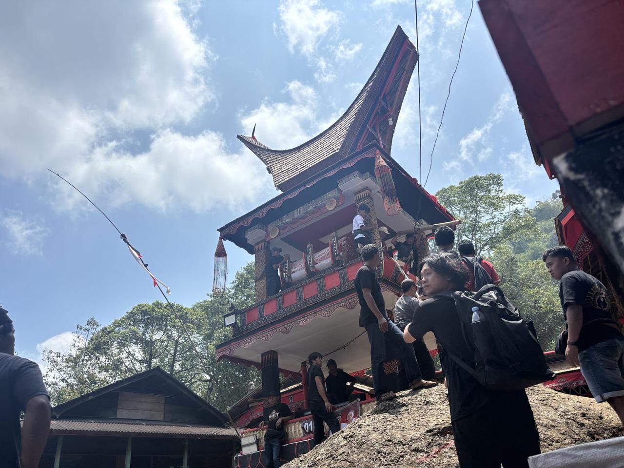 A finished tongkonan-shaped bier with a curved roof and tasseled decoration, set on top of a boulder above the plaza.