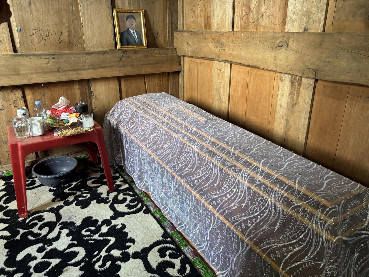 A framed portrait above a bed made up with white lace cloth, small offerings set on a red table beside it.