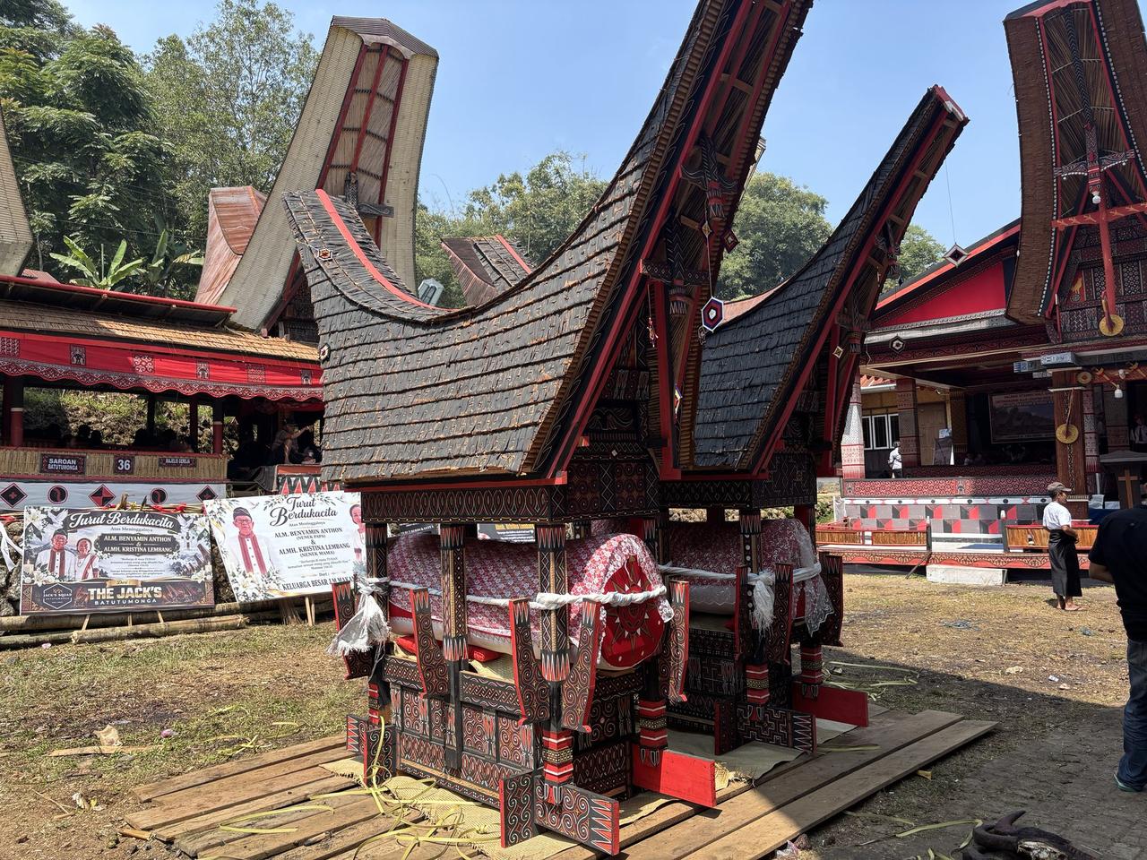 The bier set down beneath the trees, two coffins visible inside wrapped in red and white lace, condolence signboards for Benyamin Anthon and Kristina Lembang standing beside it.