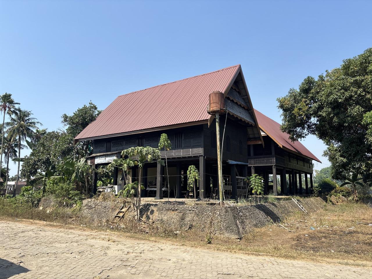 Sebuah rumah panggung tradisional beratap seng merah, terlihat dari pinggir jalan di antara pohon-pohon kelapa.
