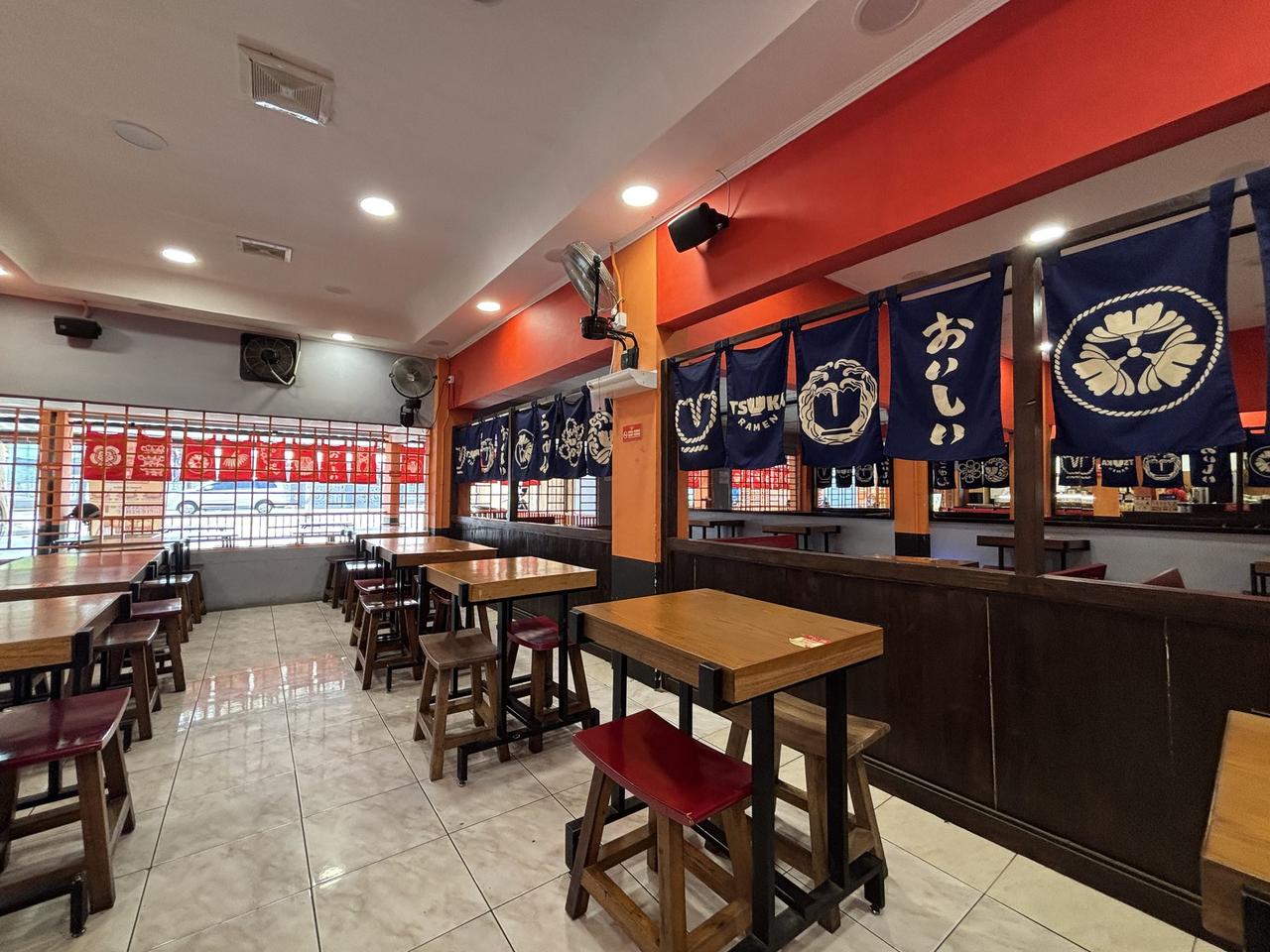 The interior dining area of a Tsuka Ramen restaurant, wooden tables and stools beneath indigo noren curtains printed with the restaurant's crest.