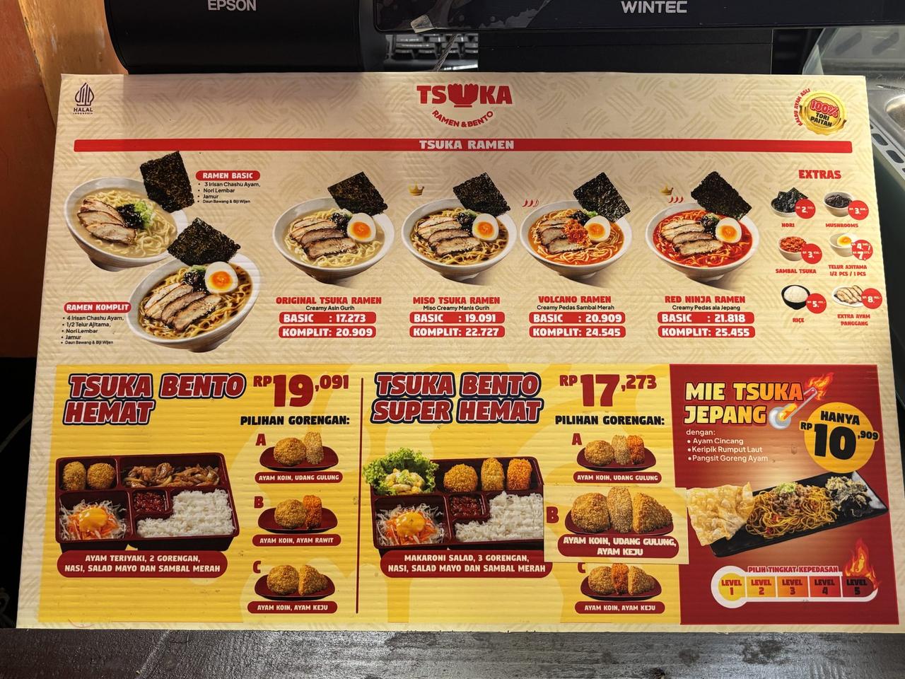 A menu board at Tsuka Ramen listing four ramen varieties with Basic and Komplit pricing, alongside bento sets and extra toppings.