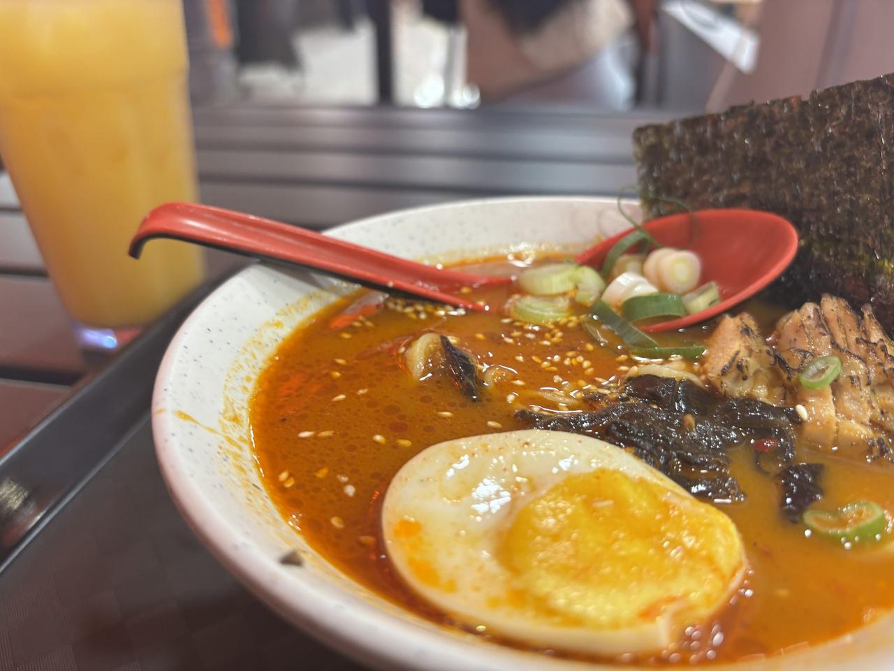 A close-up of a spicy Tsuka Ramen bowl with a red chili-infused broth, chashu, and a soft-boiled egg, served alongside a glass of orange juice.