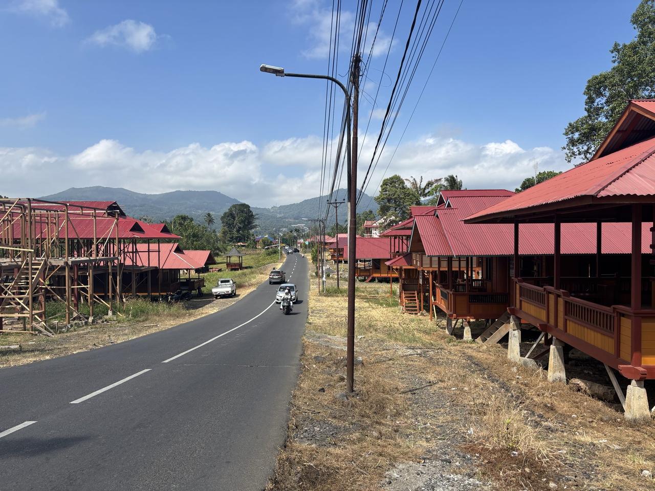 Sebuah jalan yang membentang melewati Desa Woloan, dijajari di kedua sisinya oleh rumah-rumah kayu tradisional beratap merah yang memanjang menuju perbukitan.