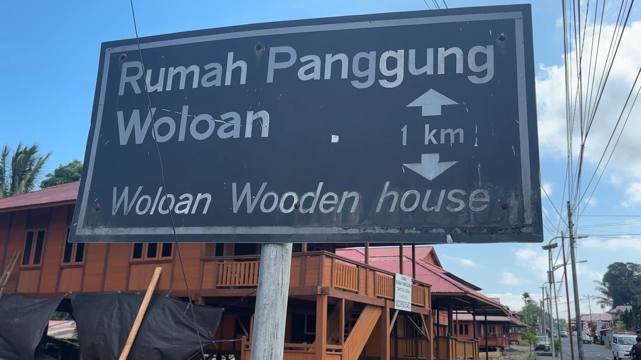 Papan penunjuk di pinggir jalan bertuliskan "Rumah Panggung Woloan / Woloan Wooden House," menunjukkan arah masuk ke desa.