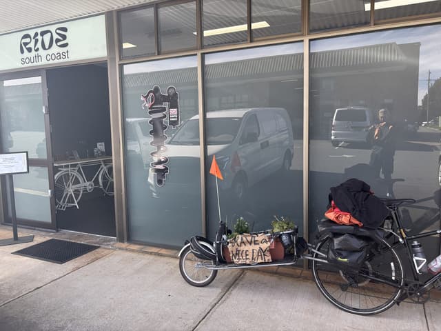 A loaded touring bicycle parked outside a bike shop, a handwritten 'have a nice day' sign tucked among potted plants on its trailer.