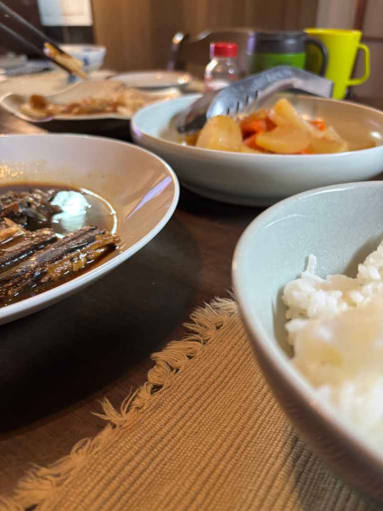A dining table featuring bowls of rice, fish in sauce, and vegetable dishes, with cutlery and drinks visible in the background.